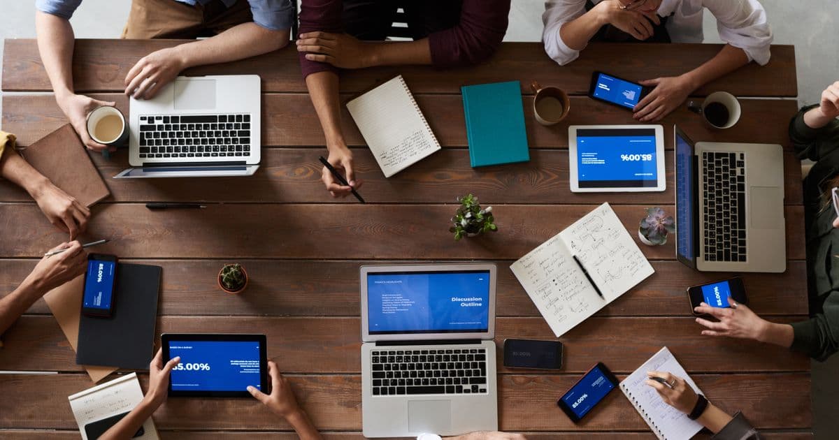 Overhead view of a team meeting table covered with laptops, tablets, and phones showing different applications.